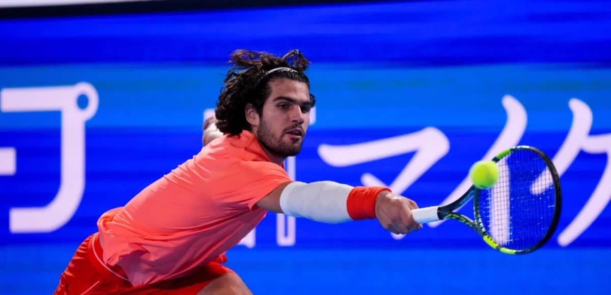 Carlos Alcaraz, con remera coral y muñequera roja, se estira para pegar una derecha frente a un cartel azul en el Japan Open 2026