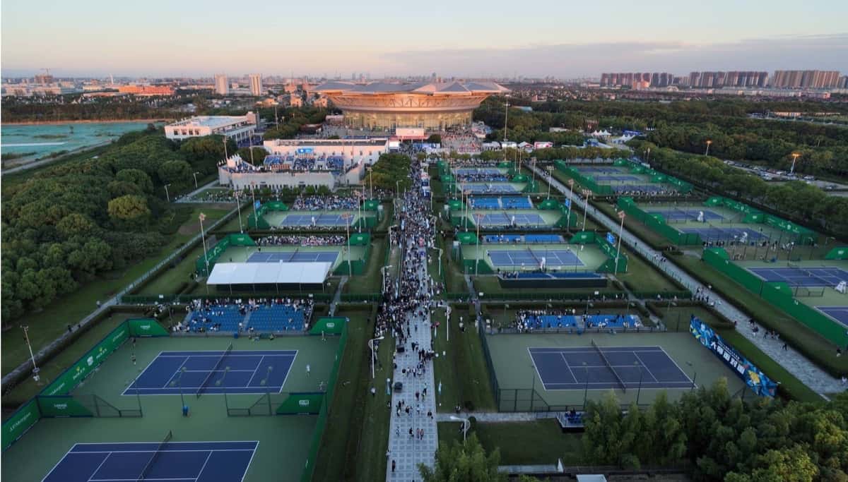 Vista aérea del Qi Zhong Tennis Center de Shanghái al atardecer, con las canchas exteriores azules y verdes, el público en el pasillo central y el estadio principal al fondo