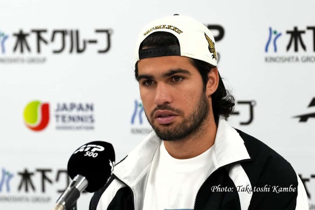 Carlos Alcaraz, con gorra blanca hacia atrás y campera blanca y negra, habla en conferencia de prensa frente al micrófono del ATP 500 en el Japan Open