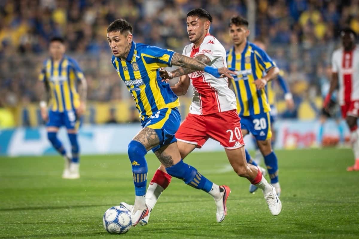 Un jugador de Rosario Central, con la camiseta azul y amarilla, conduce la pelota perseguido por un rival de camiseta blanca y roja, con la tribuna de Central detrás; foto de archivo de octubre de 2026 (foto: prensa Liga Profesional de Fútbol)