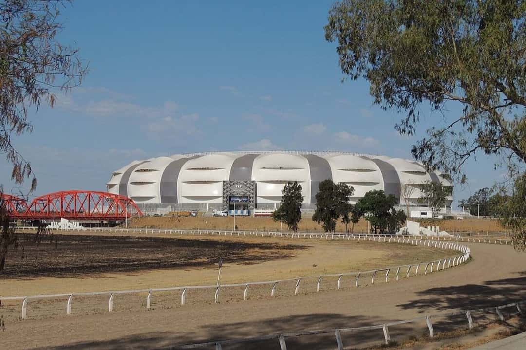 El estadio Único Madre de Ciudades, en Santiago del Estero, con su cubierta blanca y gris, visto desde el hipódromo con el puente rojo a la izquierda; foto de archivo de 2021 (foto: miroab, Wikimedia Commons, licencia CC BY-SA 2.0)