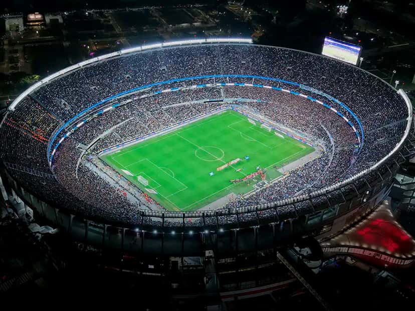 Vista aérea nocturna del Estadio Monumental con las tribunas llenas y el campo de juego iluminado, en una foto de archivo de la AFA