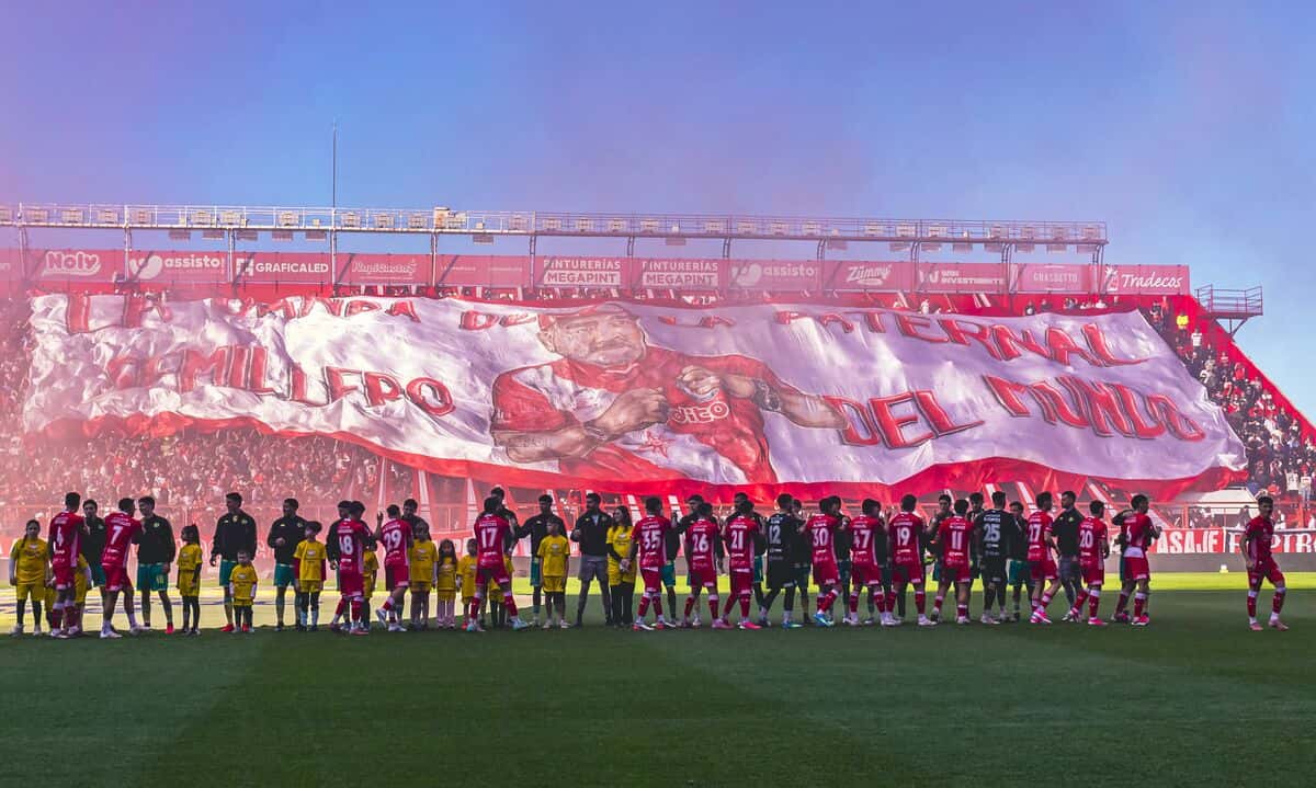 Los jugadores de Argentinos Juniors, con la camiseta roja, y los de su rival, formados en la cancha del estadio Diego Armando Maradona antes de un partido, con un telón gigante en la tribuna de fondo; foto de archivo (foto: prensa oficial de Argentinos Juniors)