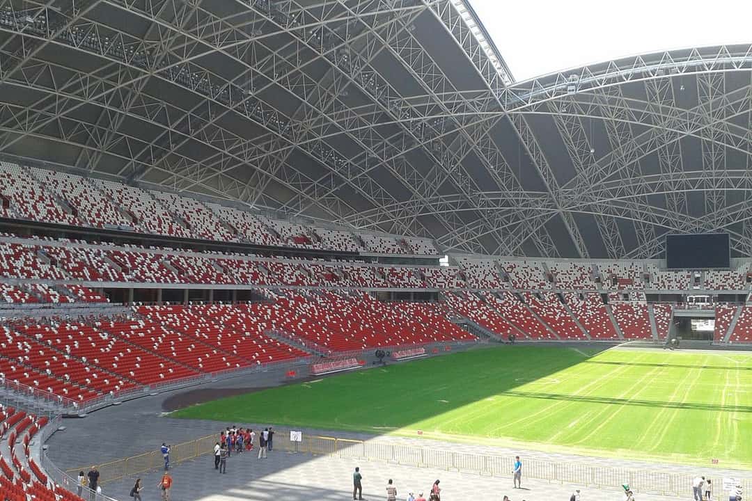 Las tribunas rojas y blancas del Estadio Nacional de Singapur, con el techo de estructura metálica y el campo de juego verde, vistas desde la platea; foto de archivo de agosto de 2014 (foto: Exec8, Wikimedia Commons, licencia CC BY-SA 4.0)