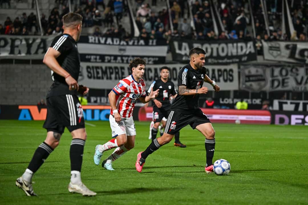 Un jugador de Deportivo Riestra, de negro, avanza con la pelota ante la marca de un futbolista de Unión con la camiseta a rayas rojas y blancas, en el estadio Guillermo Laza, durante el cierre de la fecha 13 (foto: Liga Profesional de Fútbol)