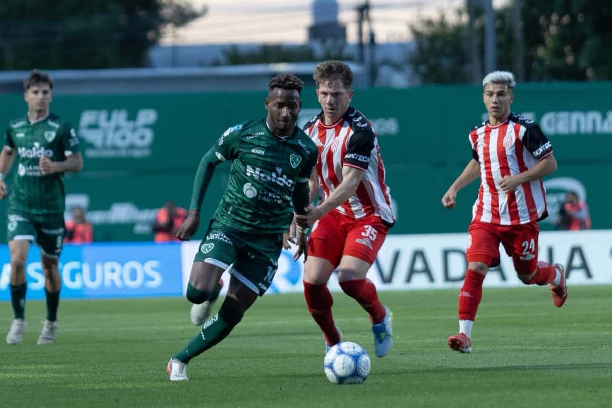 Un delantero de Sarmiento, de camiseta verde, maneja la pelota perseguido por un defensor de Barracas Central con la camiseta a rayas rojas y blancas número 35, en el estadio Eva Perón de Junín, durante la fecha 13 del Clausura (foto: Liga Profesional de Fútbol)