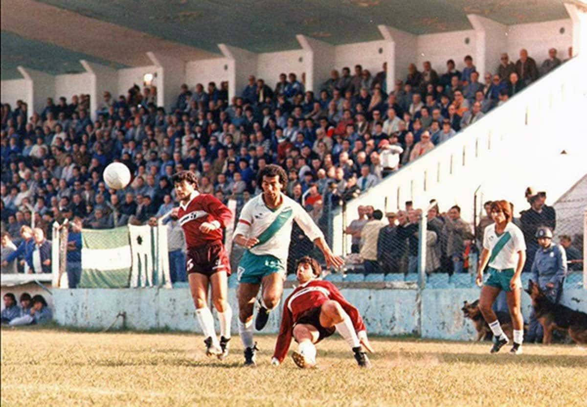 Jugadores de Banfield, con camiseta blanca y verde, y de Lanús, con camiseta granate, disputan una pelota aérea en un clásico del Sur ante una tribuna llena; foto de archivo de 1987 (foto: El Gráfico, Wikimedia Commons, dominio público)