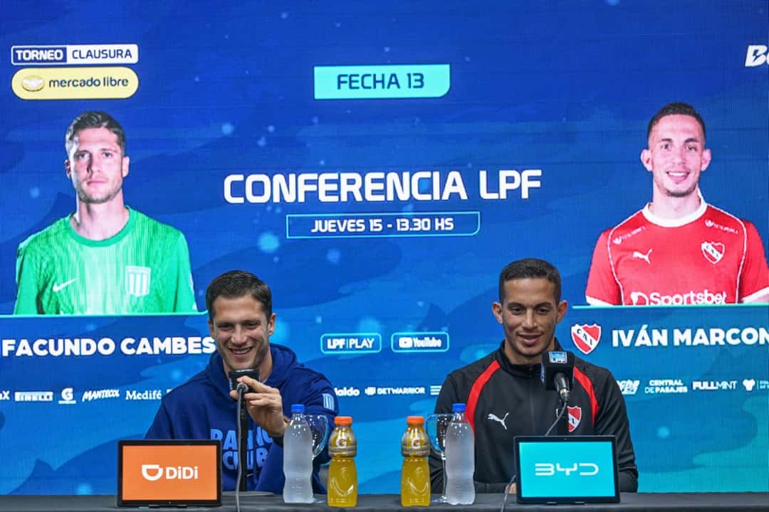 Facundo Cambeses e Iván Marcone, sonrientes, sentados frente a los micrófonos en la conferencia de la Liga Profesional por la fecha 13, con sus caras en la pantalla gigante de fondo (foto: Liga Profesional de Fútbol)