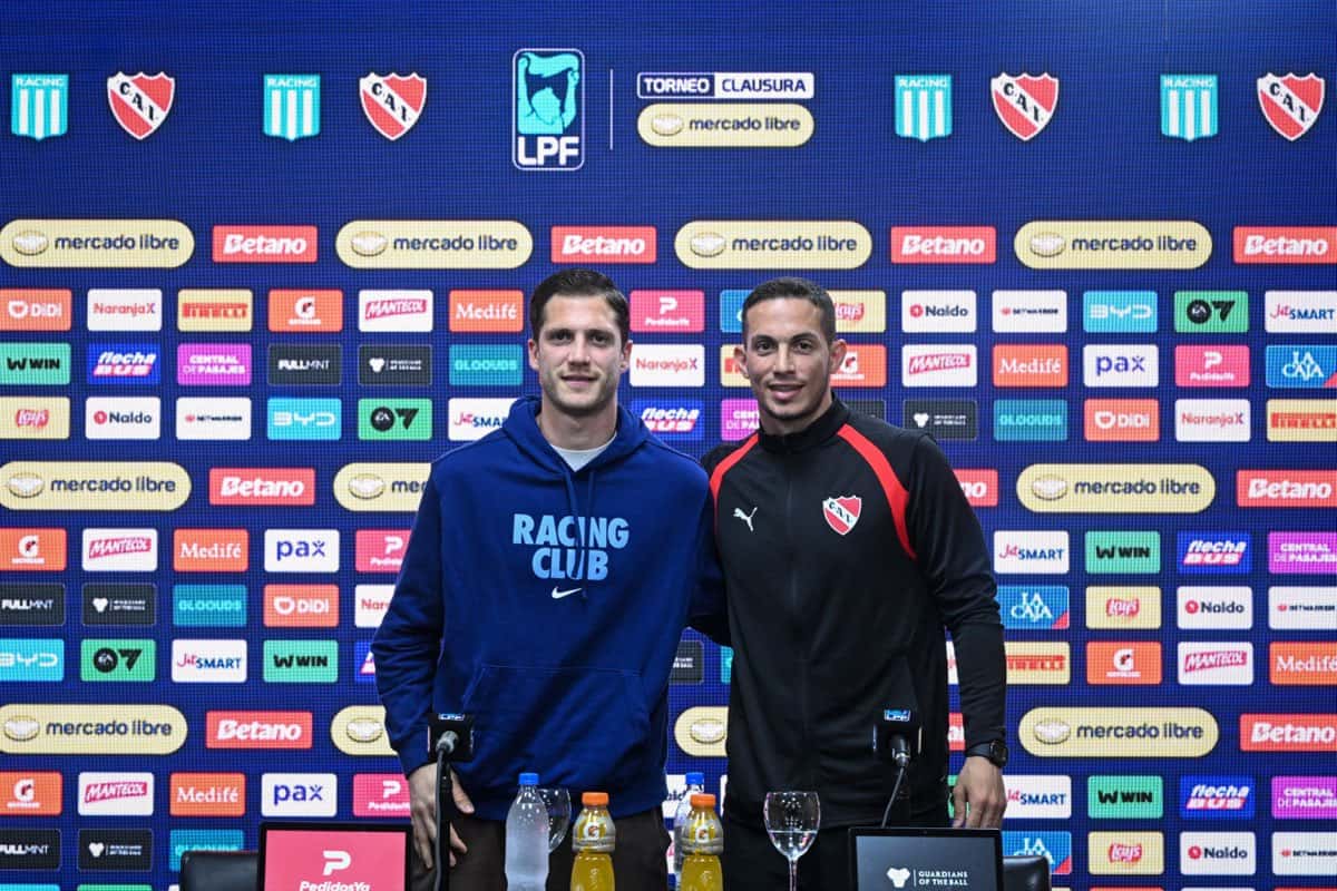 Facundo Cambeses, de Racing, con buzo azul, y Iván Marcone, de Independiente, con campera negra y roja, posan juntos frente al panel de la Liga Profesional en la conferencia previa al clásico de Avellaneda (foto: Liga Profesional de Fútbol)