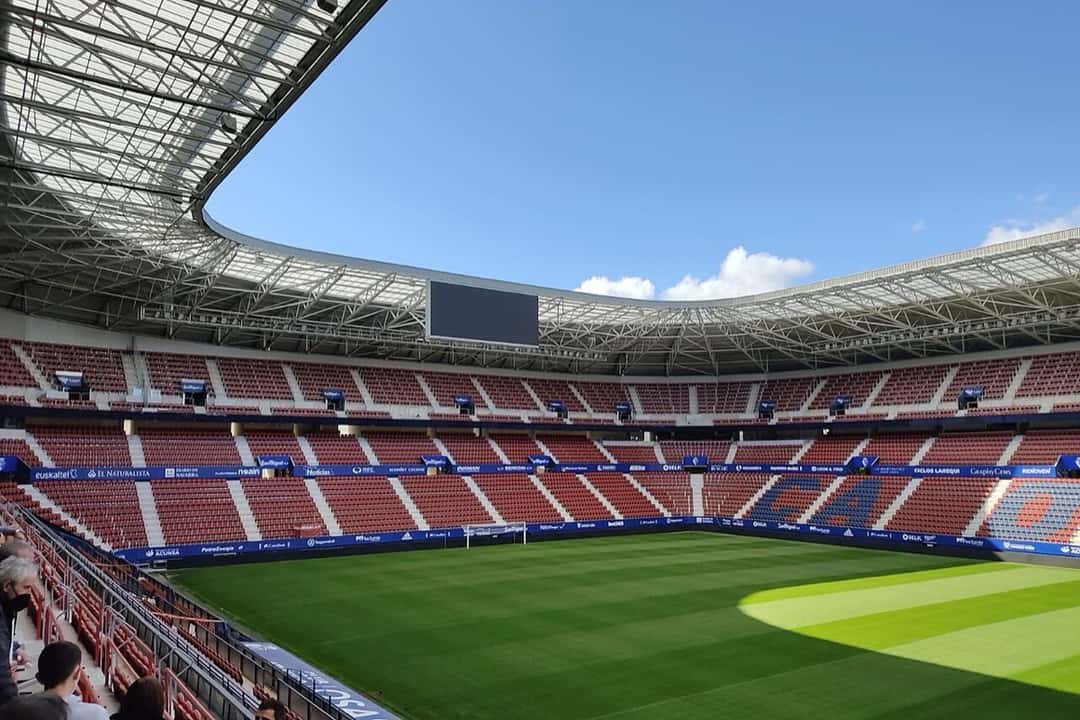 El estadio El Sadar, de Osasuna, con las butacas rojas, el techo metálico que cubre las tribunas y la cancha iluminada por el sol; foto de archivo de agosto de 2021 (foto: Visma17, Wikimedia Commons, licencia CC BY-SA 4.0)