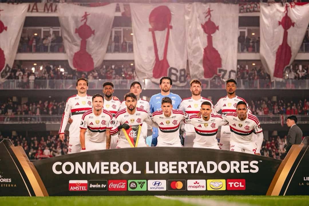 El equipo titular de Flamengo posa con su camiseta blanca detrás del cartel de la CONMEBOL Libertadores antes del partido en el estadio UNO, con las banderas de Estudiantes colgadas en la tribuna (foto: Adriano Fontes / prensa oficial de Flamengo, CRF)