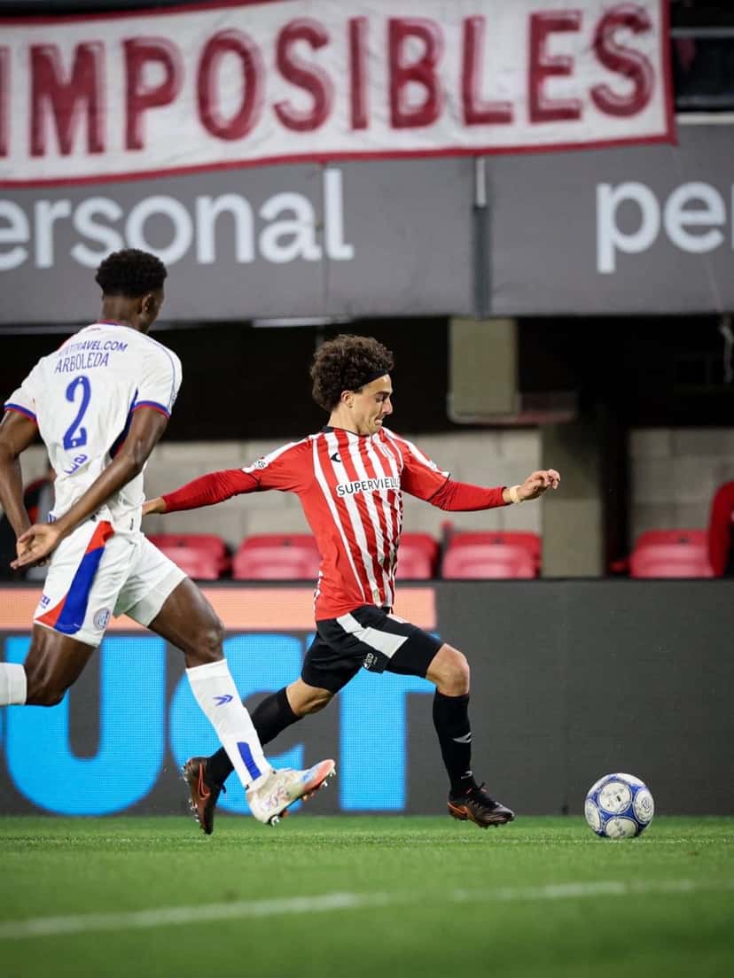 Un jugador de Estudiantes, con la camiseta roja y blanca, conduce la pelota perseguido por un jugador de San Lorenzo con el número 2, en un partido de noche en el estadio UNO (foto: Liga Profesional de Fútbol)