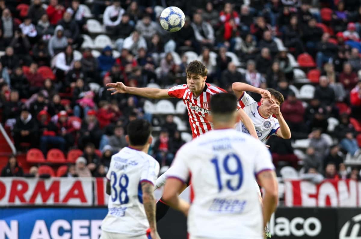 Un jugador de Estudiantes, con la camiseta roja y blanca, salta a cabecear entre dos jugadores de San Lorenzo, de blanco, con los números 38 y 19, frente a la tribuna del estadio UNO (foto: prensa oficial de Estudiantes de La Plata)