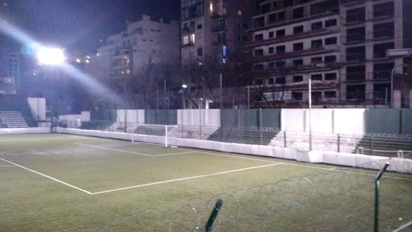 La tribuna de Excursionistas iluminada de noche, con la cancha vacía y los edificios del barrio detrás; foto de archivo de 2016 (foto: EstudiantesFotografias, Wikimedia Commons, licencia CC BY-SA 4.0)
