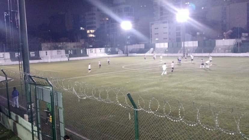 Un partido nocturno en la cancha de Excursionistas, con las luces encendidas y el nombre del club pintado en el paredón del fondo; foto de archivo de 2016 (foto: EstudiantesFotografias, Wikimedia Commons, licencia CC BY-SA 4.0)