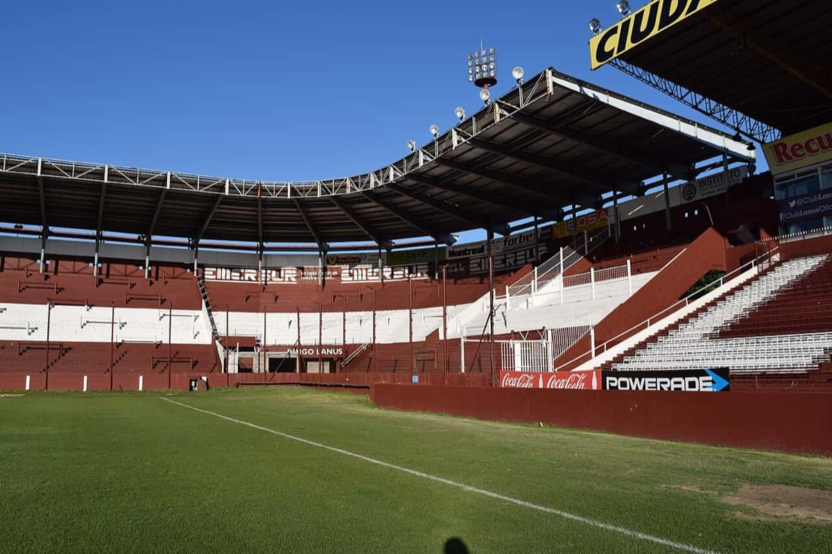 La platea y la tribuna norte del estadio Ciudad de Lanús, con las gradas granates y blancas vacías y el campo de juego en primer plano (foto de archivo de 2014: Mauricio V. Genta, Wikimedia Commons, CC0)