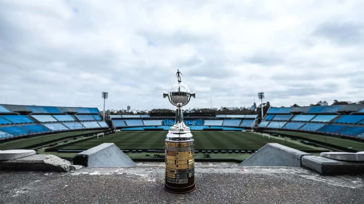 El trofeo de la CONMEBOL Libertadores sobre una base de cemento, con la cancha y las tribunas celestes del Estadio Centenario de Montevideo de fondo (foto: prensa Estudiantes de La Plata)