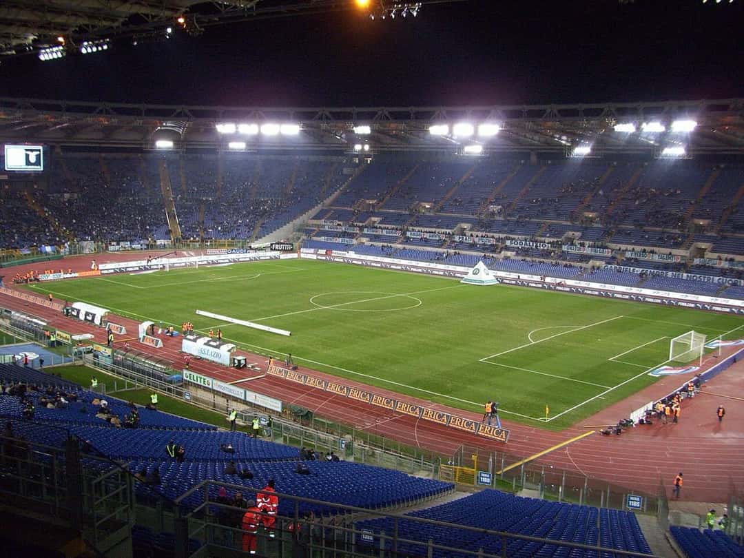 El Estadio Olímpico de Roma con la cancha iluminada y las tribunas azules, durante un partido nocturno de la Lazio; foto de archivo de 2009 (foto: Alexdevil, Wikimedia Commons, licencia CC BY-SA 3.0)
