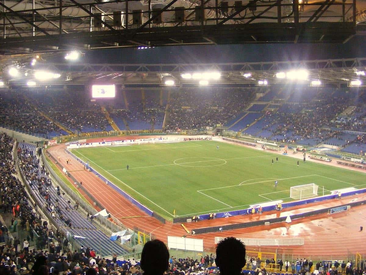 El Estadio Olímpico de Roma iluminado de noche durante un partido de la Lazio, con las tribunas azules y la pista de atletismo alrededor de la cancha; foto de archivo de 2008 (foto: Andrew, Wikimedia Commons, licencia CC BY-SA 3.0)