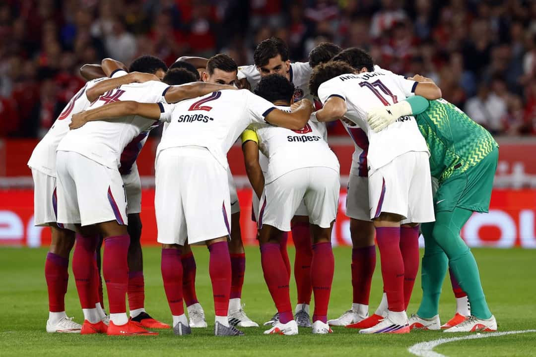 Los jugadores del PSG, con la camiseta blanca y las medias rojas, se abrazan en ronda en la cancha antes de un partido de la Ligue 1, con la tribuna detrás; foto de archivo (foto: prensa Paris Saint-Germain)