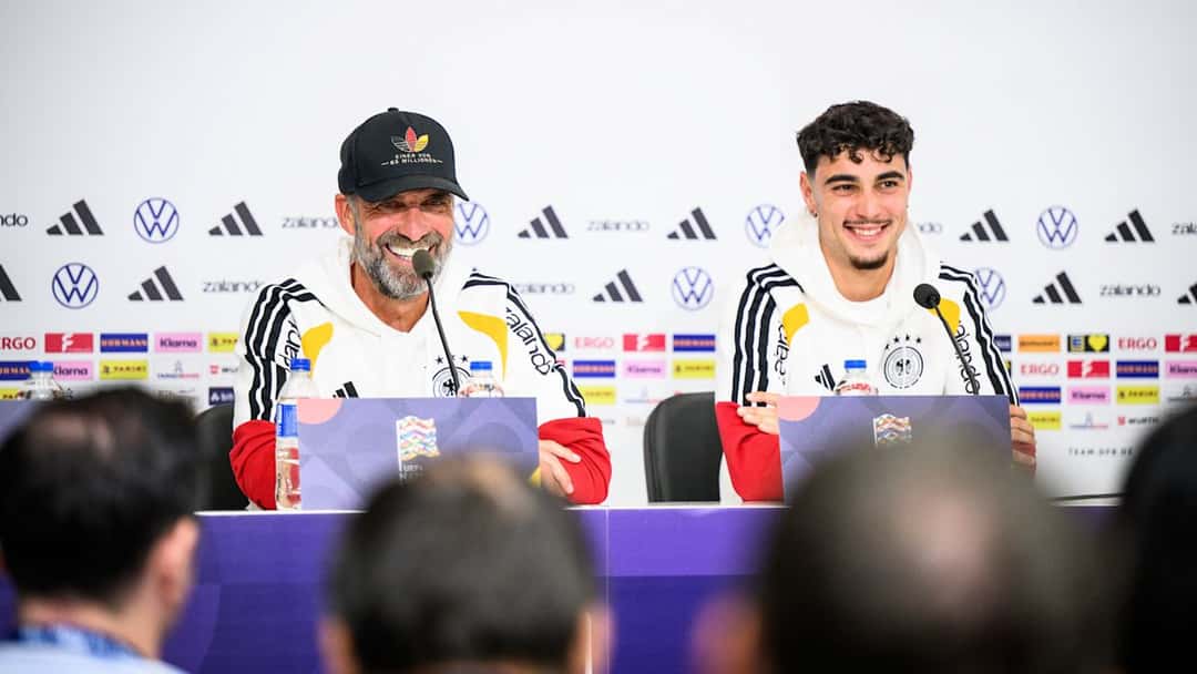 Jürgen Klopp, con gorra negra, y Aleksandar Pavlović sonríen en la conferencia de prensa de la selección alemana, frente a un panel de patrocinadores
