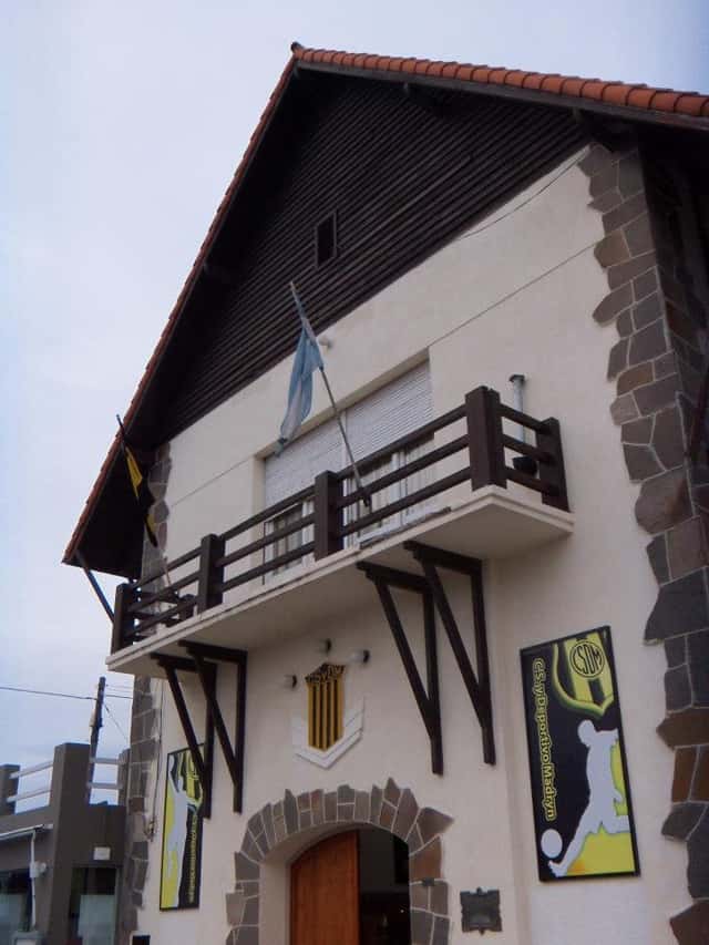 La sede del Club Social y Deportivo Madryn, con su fachada blanca, piedra y balcón de madera; foto de archivo de 2018 (foto: Ulises Icardi, Wikimedia Commons, licencia CC BY-SA 4.0)