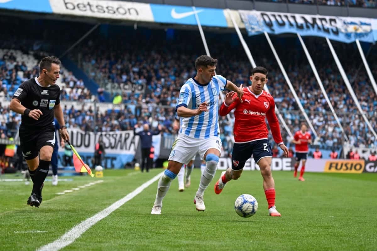 Un jugador de Racing, con la camiseta celeste y blanca, maneja la pelota perseguido por un jugador de Independiente, de rojo, junto a la línea lateral del Cilindro de Avellaneda, con el árbitro a la izquierda (foto: Liga Profesional de Fútbol)