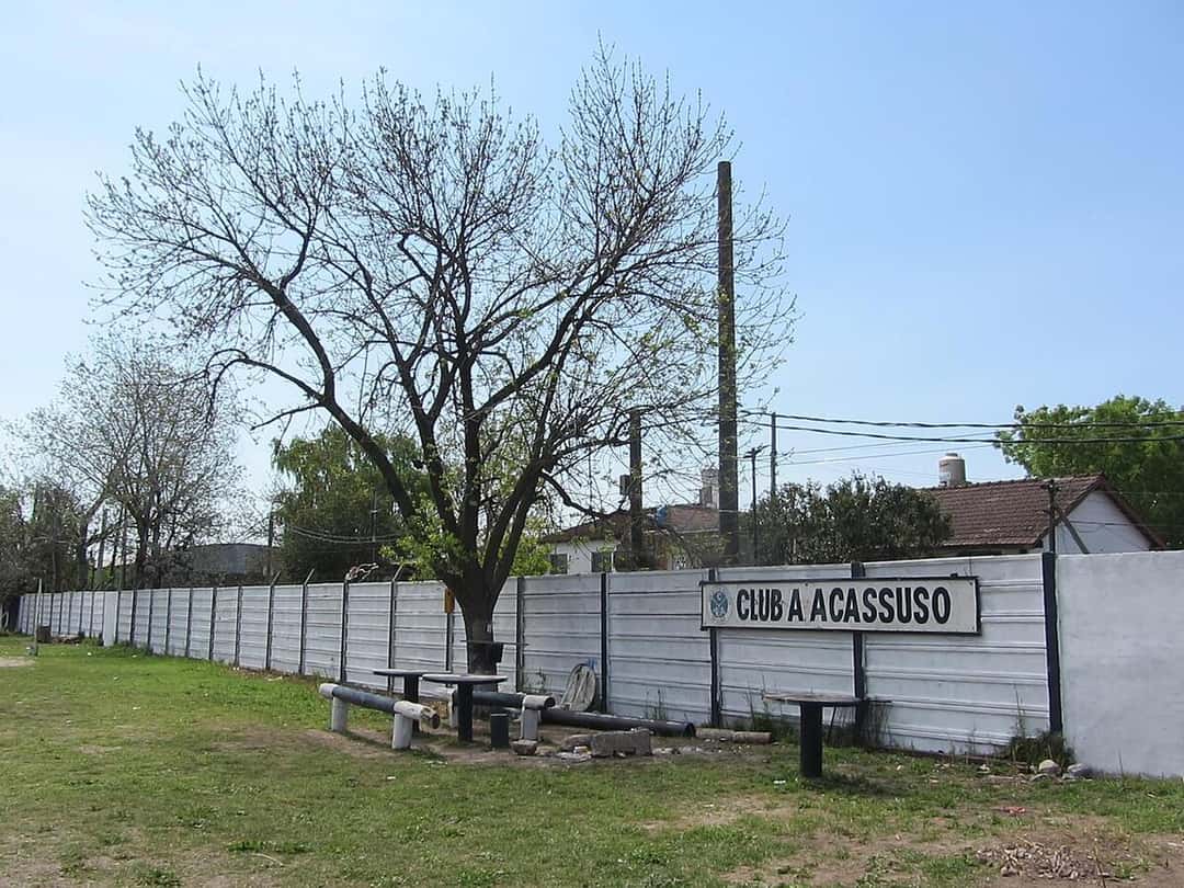 La entrada del estadio de Acassuso, con un paredón blanco, un cartel con el nombre del club y árboles en la vereda; foto de archivo de 2021 (foto: Ulises Icardi, Wikimedia Commons, licencia CC BY-SA 4.0)