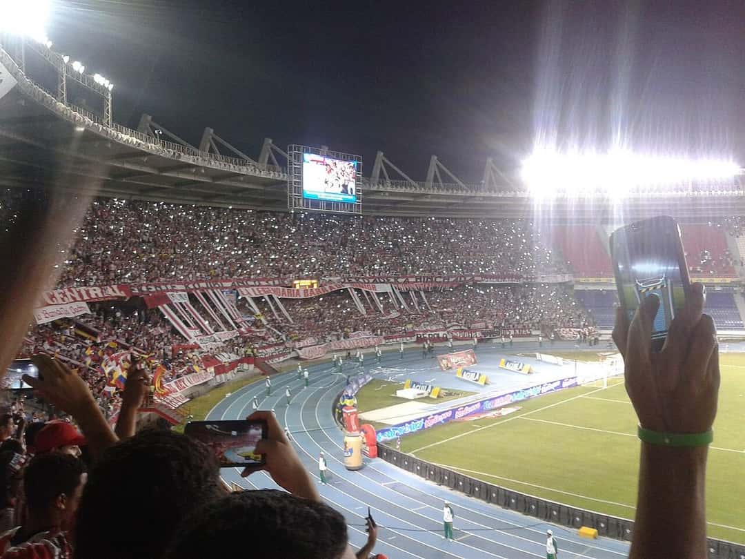 Hinchas en la tribuna del estadio Metropolitano de Barranquilla, de noche, con la pista de atletismo y la cancha de fondo; foto de archivo de 2015 (foto: Jeisonbeta, Wikimedia Commons, licencia CC BY-SA 4.0)