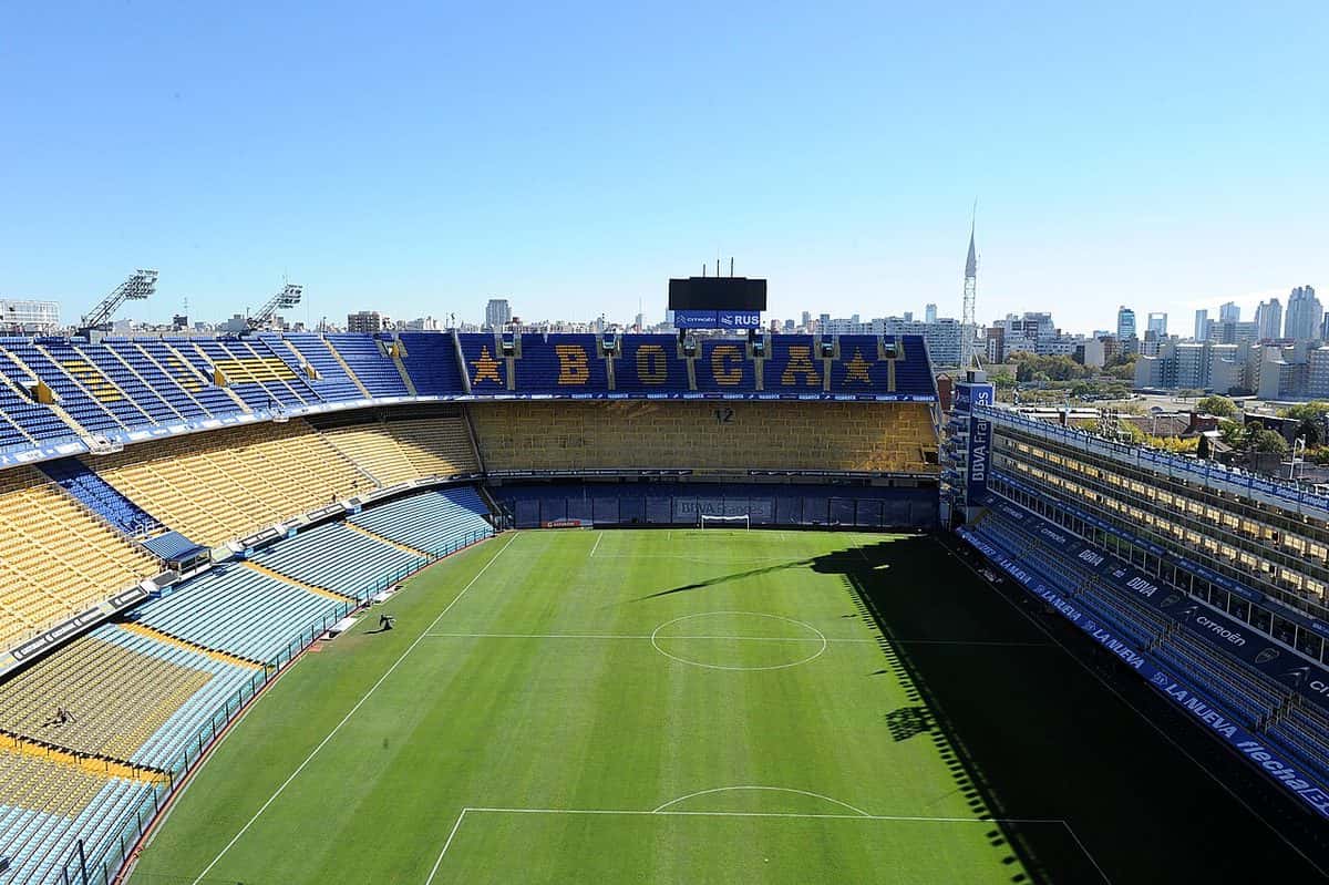 La Bombonera vacía y a pleno sol, con las tribunas azules y amarillas, la palabra BOCA escrita en los asientos y los edificios de la ciudad de fondo; foto de archivo de mayo de 2015 (foto: Prensa TV Pública, Wikimedia Commons, licencia CC BY 2.0)