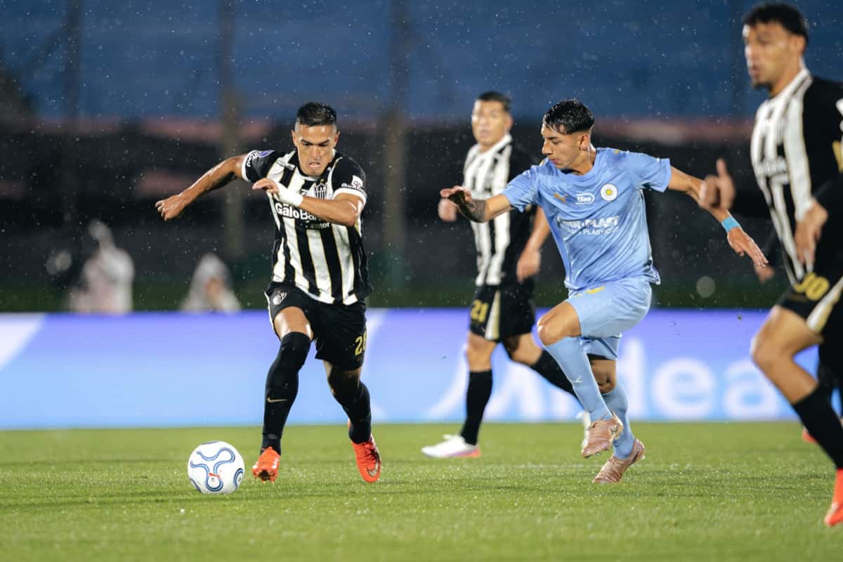 Un jugador de Atlético Mineiro, con la camiseta a rayas blancas y negras, conduce la pelota en el césped mojado del Centenario ante la marca de un futbolista de Montevideo City Torque, de celeste (foto: Pedro Souza / prensa oficial de Atlético Mineiro)