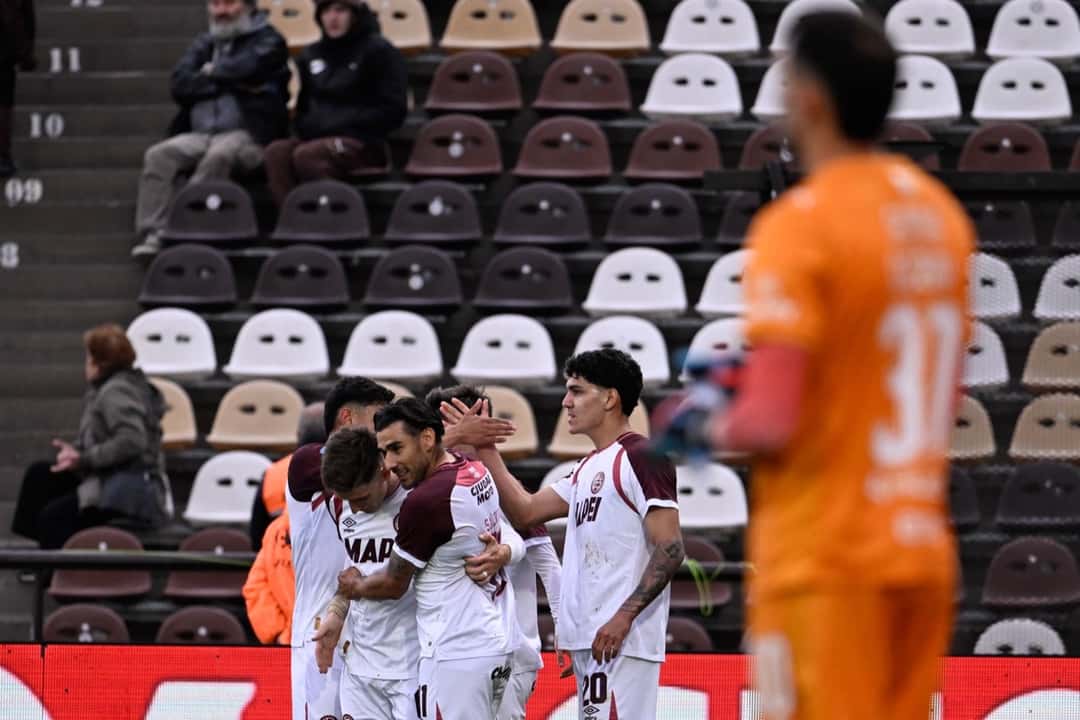 Jugadores de Lanús, de blanco y bordó, se abrazan tras el gol ante Platense, con el arquero local, de naranja, fuera de foco en primer plano (foto: Liga Profesional de Fútbol)