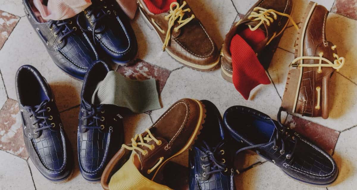 Varios pares del Timberland x Jacquemus Authentic Boat Shoe sobre un piso de baldosas claras y bordó: los navy con cuero de relieve de cocodrilo y cordones azules, y los dark brown con puntera de gamuza marrón, cordones amarillo claro y medias rojas, verdes y amarillas asomando de los zapatos