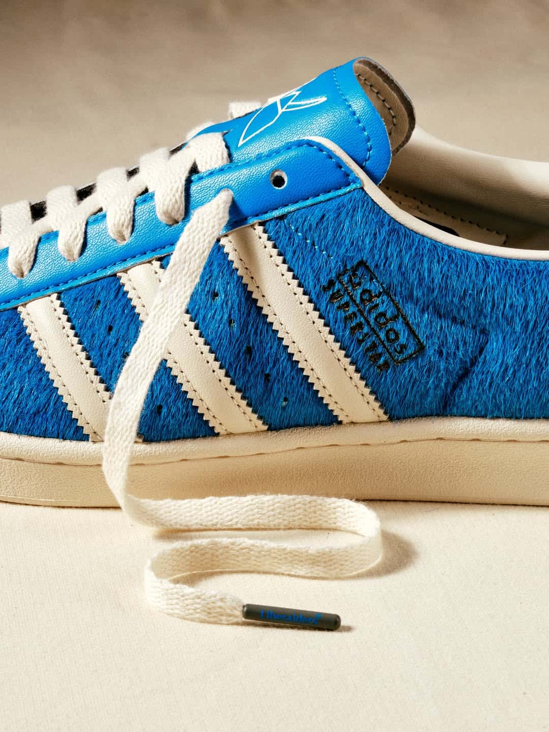 Lateral de la adidas Superstar de Liberaiders con el pelo azul en primer plano, las tres tiras crema, el logo adidas Superstar en el costado y un cordón suelto con el herrete gris que dice Liberaiders