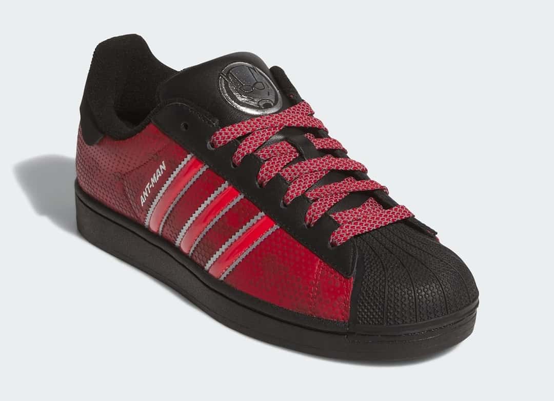 adidas Superstar Ant-Man en vista de tres cuartos con laterales rojos texturados, tiras rojas con bordes plateados, puntera negra y el logo plateado del personaje en la lengüeta