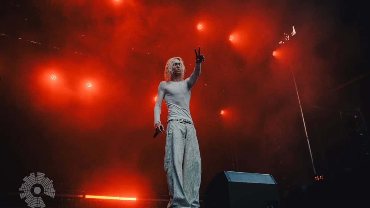 Imagen del anuncio oficial de «The Pirouette Tour»: 2hollis de pelo largo y rubio, con remera gris y pantalón ancho, levanta la mano en un escenario bañado de luces rojas