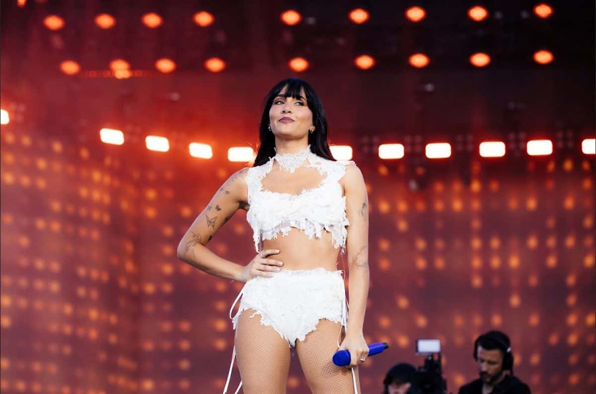 Aitana en el escenario del Cuarto Azul World Tour, con flequillo, conjunto blanco de dos piezas con flecos y un micrófono azul en la mano, frente a una pared de luces anaranjadas (foto de archivo de la gira)