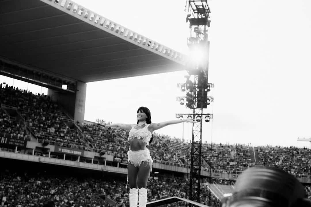 Aitana en blanco y negro, con los brazos abiertos sobre una pasarela, frente a las tribunas llenas de un estadio, con una torre de luces encendida a la derecha (foto de archivo de la gira)