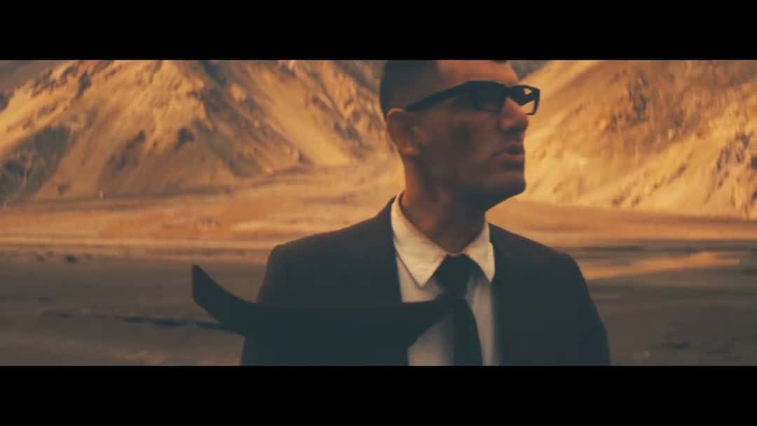 Alex Anwandter con anteojos oscuros, traje y corbata, en un paisaje de cerros áridos, en un fotograma del video oficial de «Cordillera» (foto de archivo)