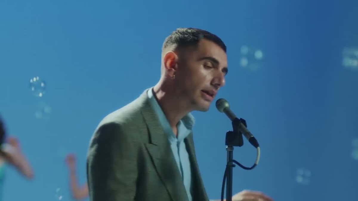 Alex Anwandter canta frente a un micrófono de pie, con saco verde claro, sobre un fondo azul con burbujas, en un fotograma del video oficial de «Maricoteca» (foto de archivo)