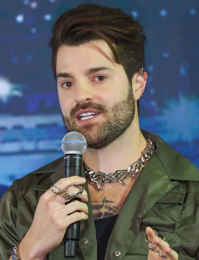 Alok, en una foto de archivo de marzo de 2024, con el pelo peinado hacia atrás, barba, campera verde y cadena plateada al cuello, hablando con un micrófono en la mano durante una conferencia en Brasilia. Foto: Renato Alves/Agência Brasília, Wikimedia Commons, CC BY 2.0