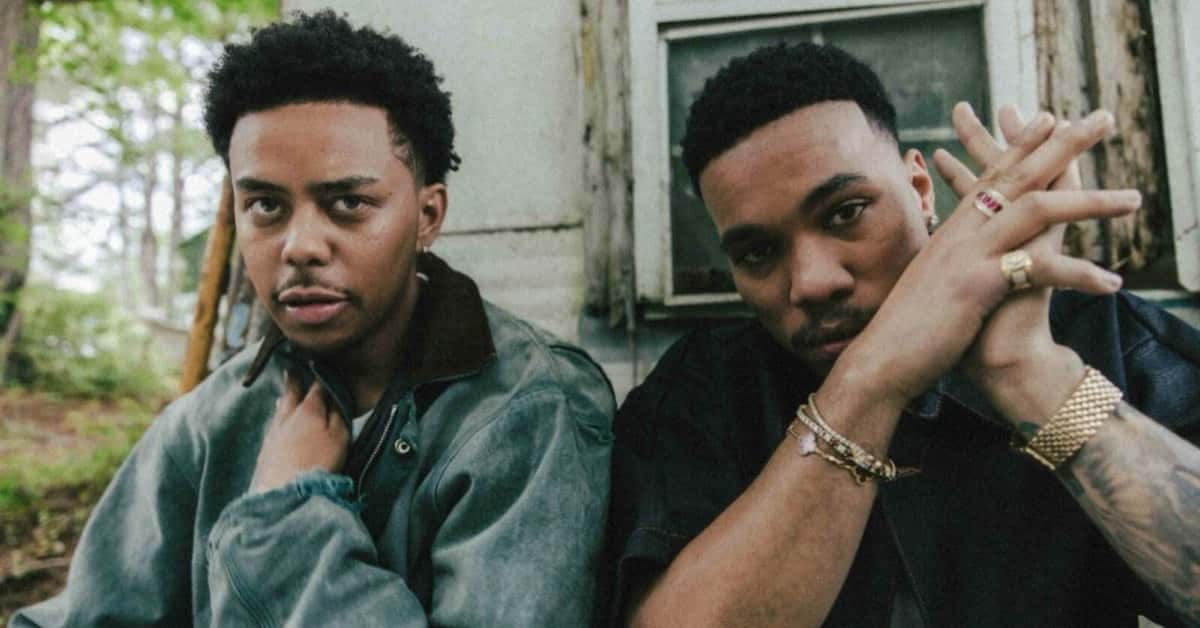Cordae con campera de jean gastada y Anderson .Paak con camisa oscura, anillos y pulseras doradas y las manos entrelazadas, sentados frente a la pared despintada de una casa de madera