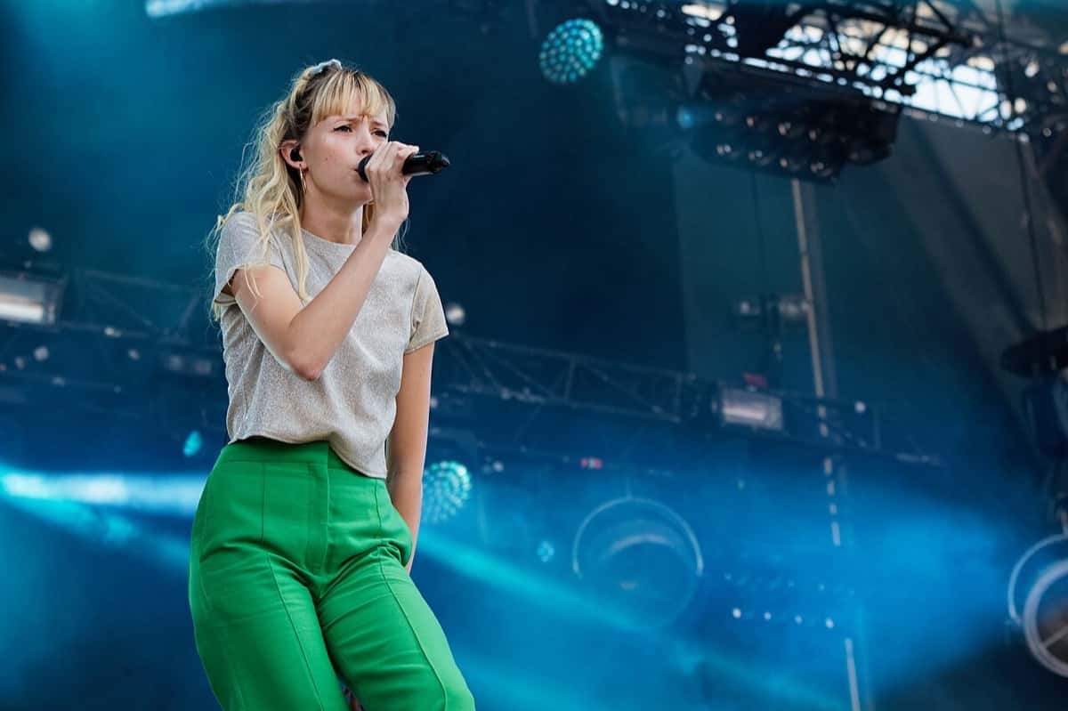 Angèle canta con el micrófono en la mano en un escenario del festival Vieilles Charrues de 2018, con remera gris brillante y pantalón verde, frente a una estructura de luces azules (foto de archivo)