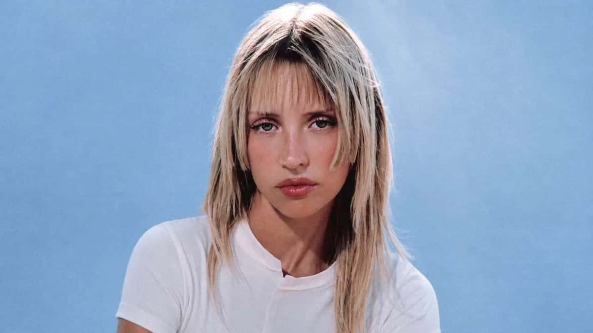 Retrato de prensa de Angèle de frente, con el pelo rubio en capas y flequillo, remera blanca y fondo celeste. Foto: Kenza Le Bas