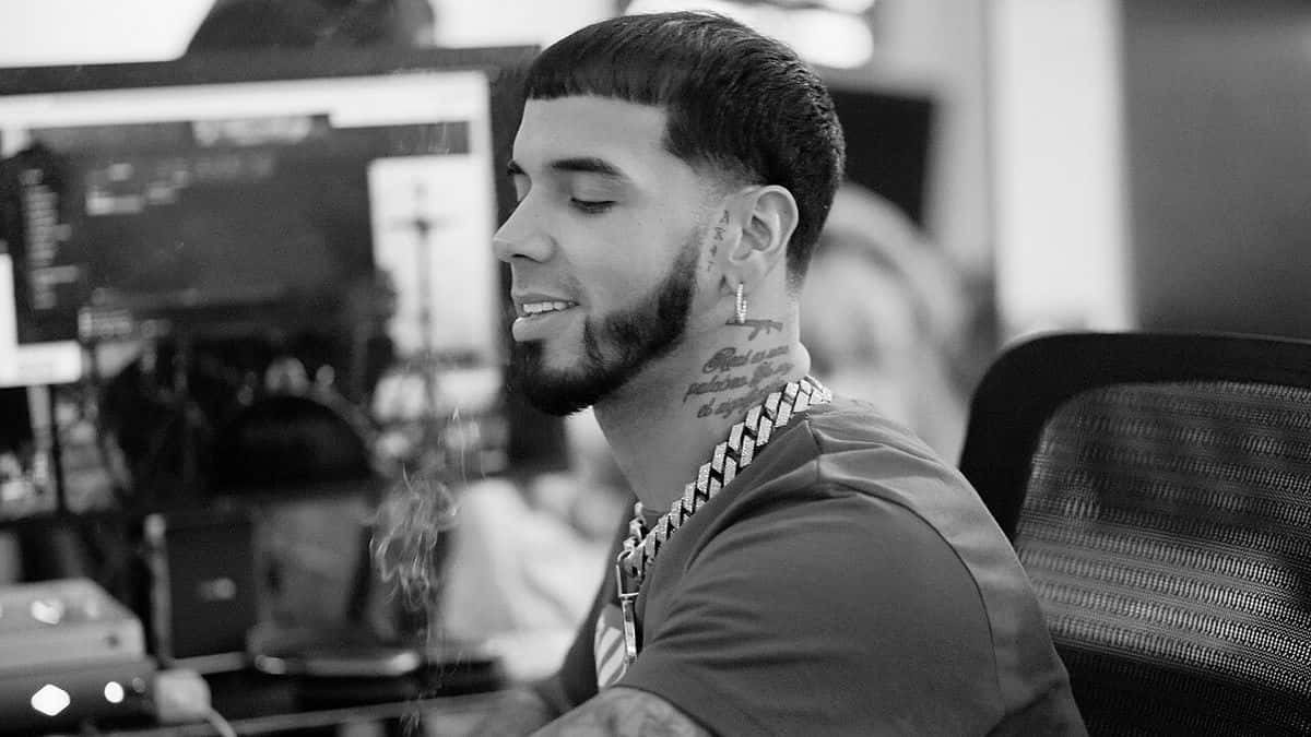Foto de archivo de Anuel AA en 2022, en blanco y negro: de perfil y sonriendo, con cadenas gruesas y tatuajes en el cuello, sentado en un estudio de grabación frente a una consola y un monitor. Foto: Timelezzzz, Wikimedia Commons, CC BY-SA 4.0