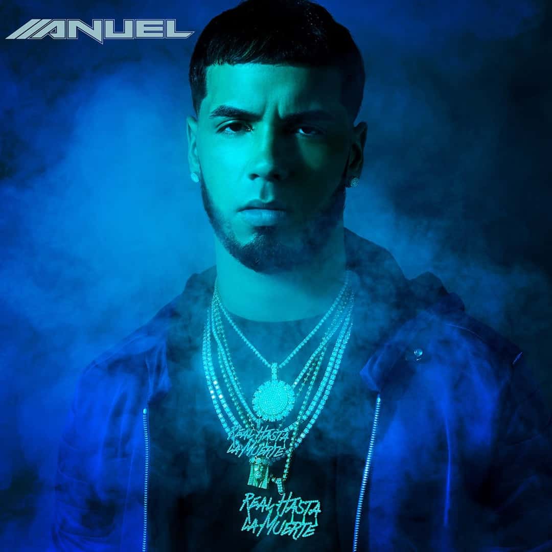 Portada de «Real Hasta la Muerte» (2018), el disco debut de Anuel AA: él, con cadenas y campera oscura, mira a cámara entre humo y luz celeste, con su logo arriba a la izquierda