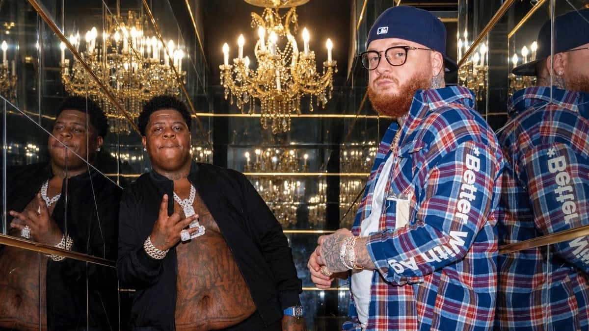 BigXthaPlug, con campera negra y cadena plateada, y Belly Gang Kushington, de gorra azul, anteojos y camisa escocesa, junto a otro acompañante, en un pasillo de espejos con arañas de cristal, en la imagen oficial de "LITE"
