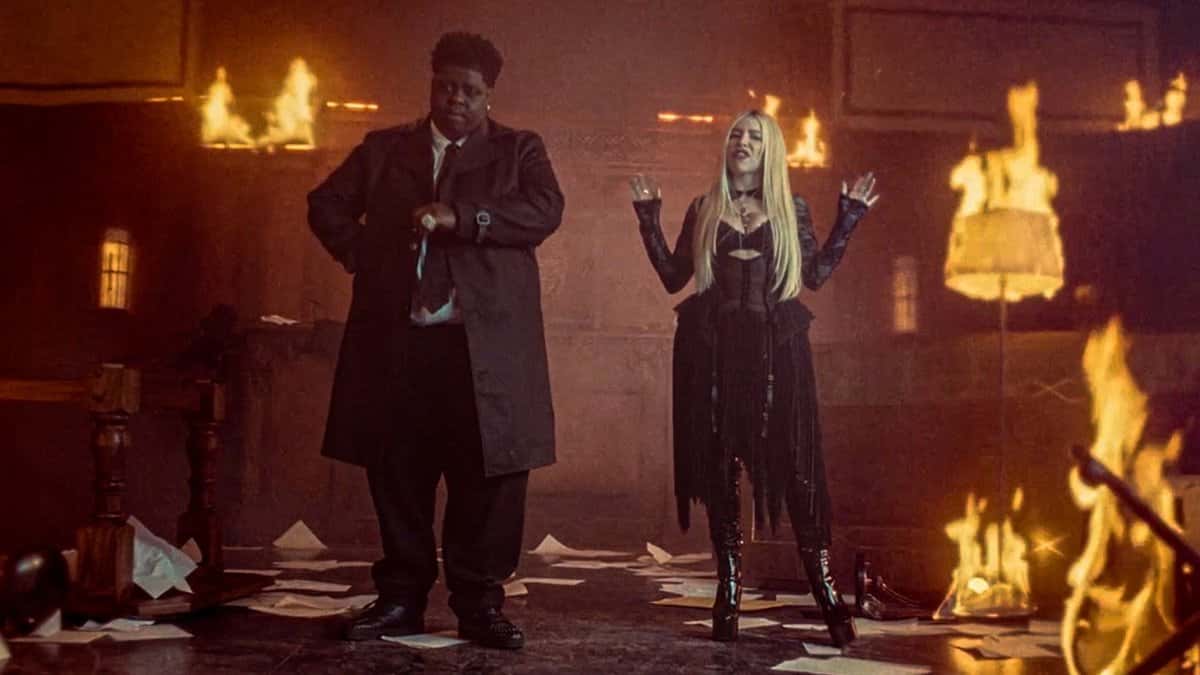 BigXthaPlug, con sobretodo negro, y Ava Max, de pelo rubio largo y ropa negra, en una sala con lámparas y muebles prendidos fuego, en una imagen del video oficial de «BURN»
