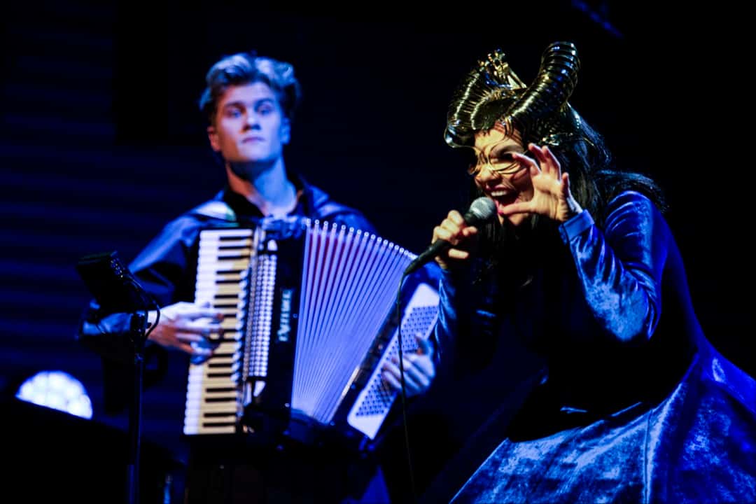 Foto oficial del concierto Orkestral con el coro Hamrahlíð: Björk, con un tocado dorado con forma de cuernos, canta con gesto teatral mientras un músico la acompaña en acordeón