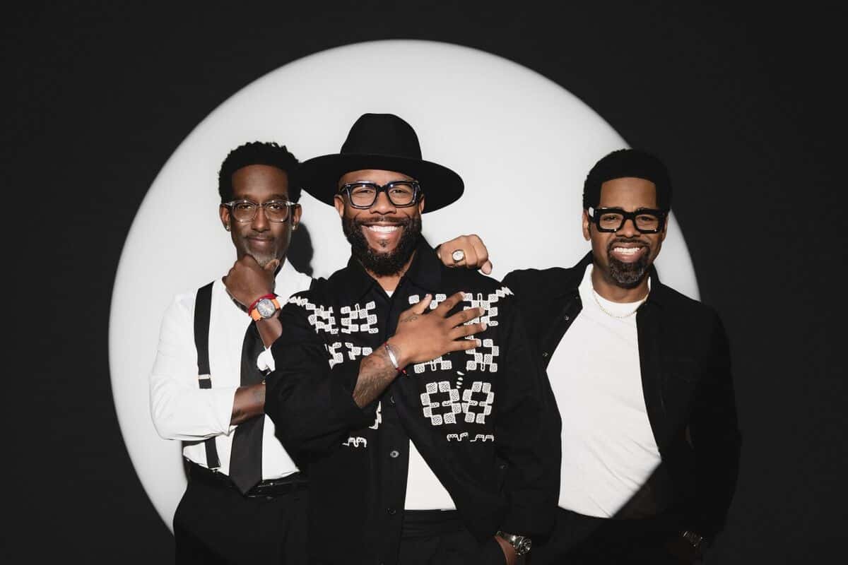 Foto de prensa de Boyz II Men: los tres integrantes posan sonrientes, con anteojos y uno con sombrero negro, frente a un círculo de luz blanca sobre fondo negro